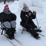 Two smiling women in a wintry landscape on sit skis.
